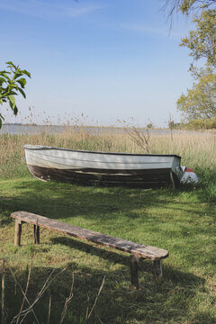 Old Wooden Rowboat On Grass By Lake With Rustic Bench And Reeds In Summer